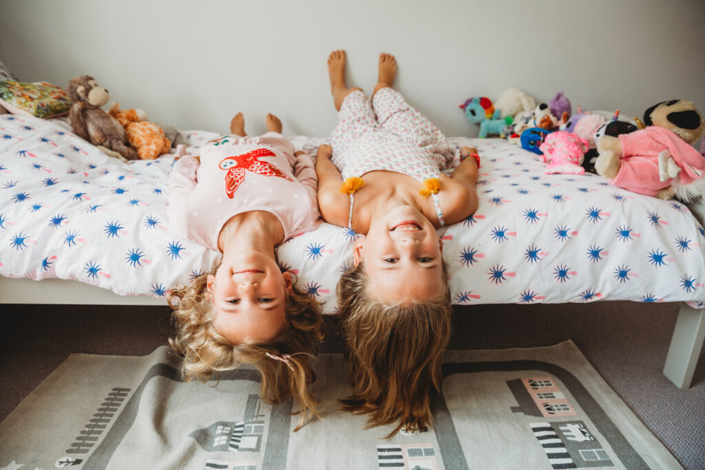 2 girls lying in bed hanging their heads down and smiling.