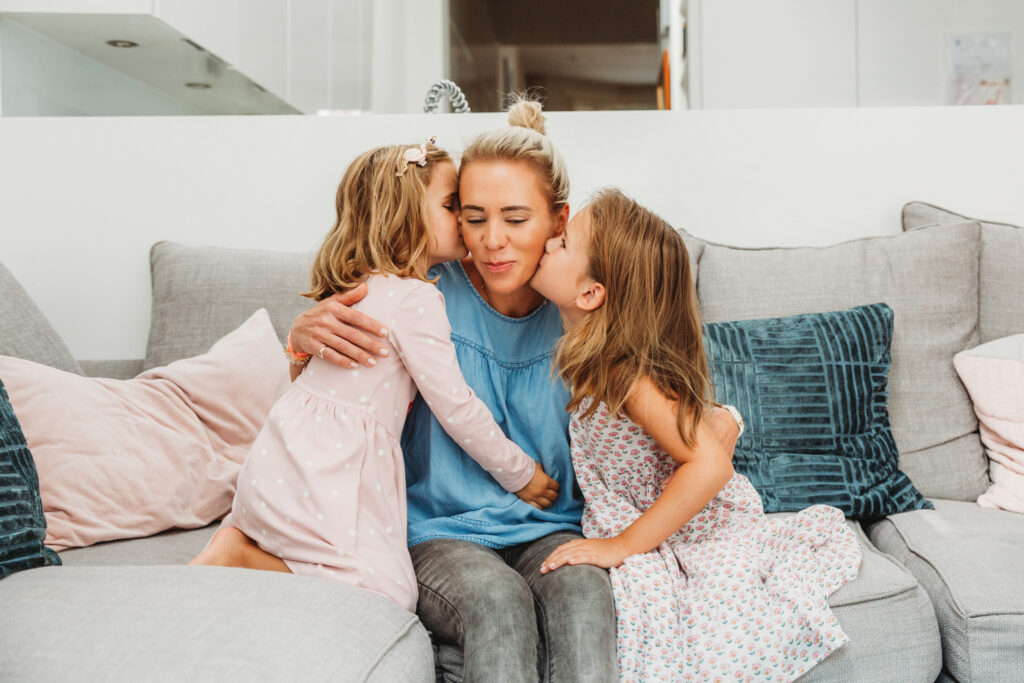 Mum sitting down on the sofa being kissed by her 2 little girls.