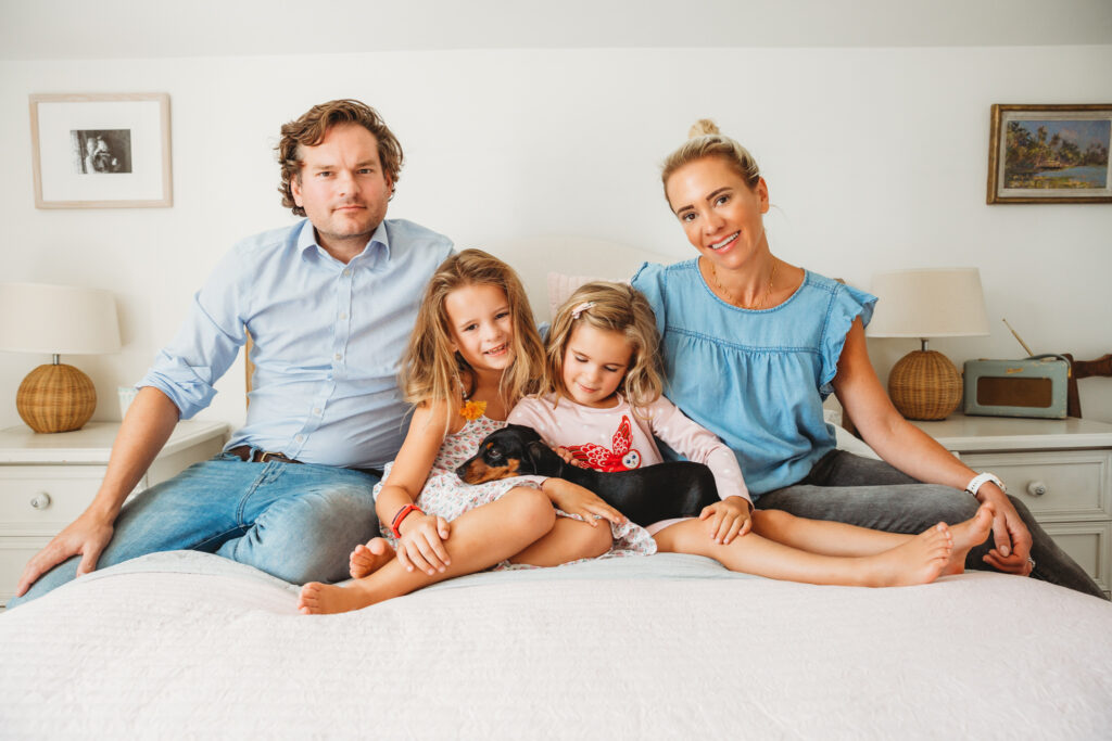 All family posing on bed with their 2 little girls in the middle holding their little puppy.