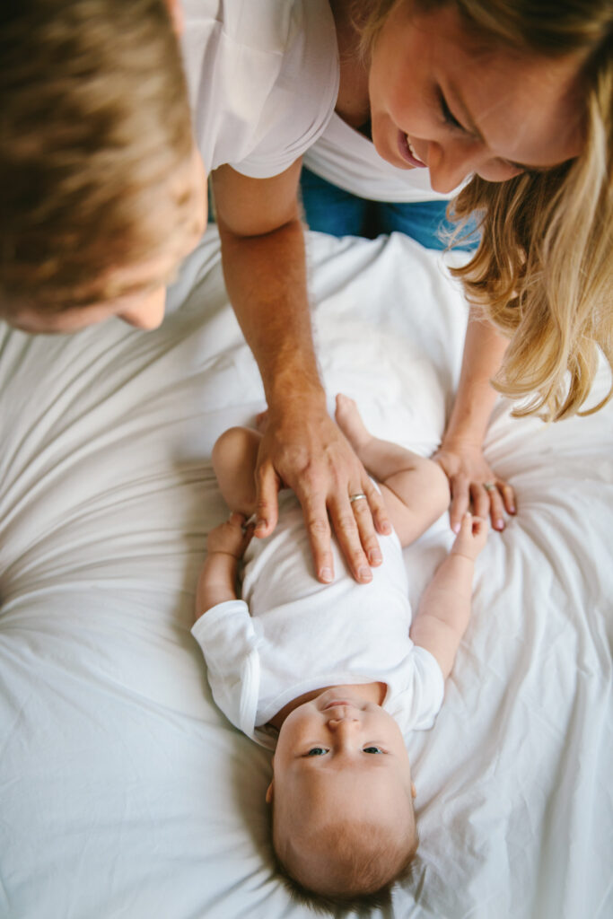 A baby lying in bed being teased by his parents who are happy to have him.