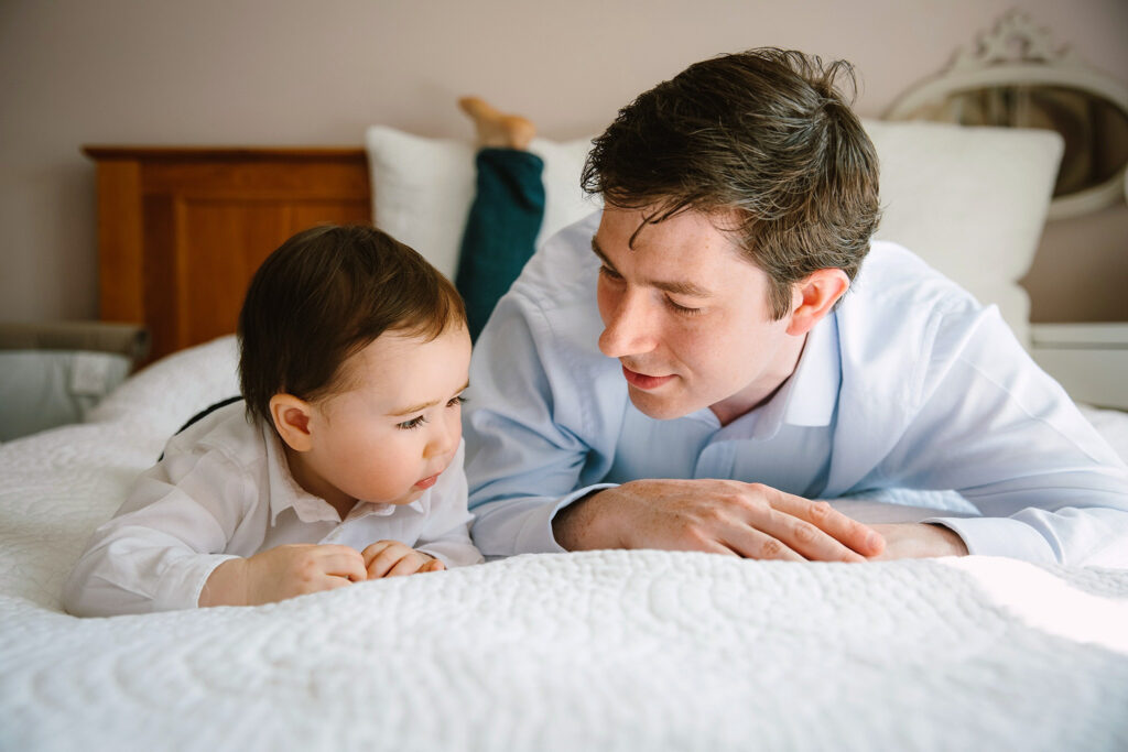 Dad and toddler lying on bed on their tummies talking to each other.
