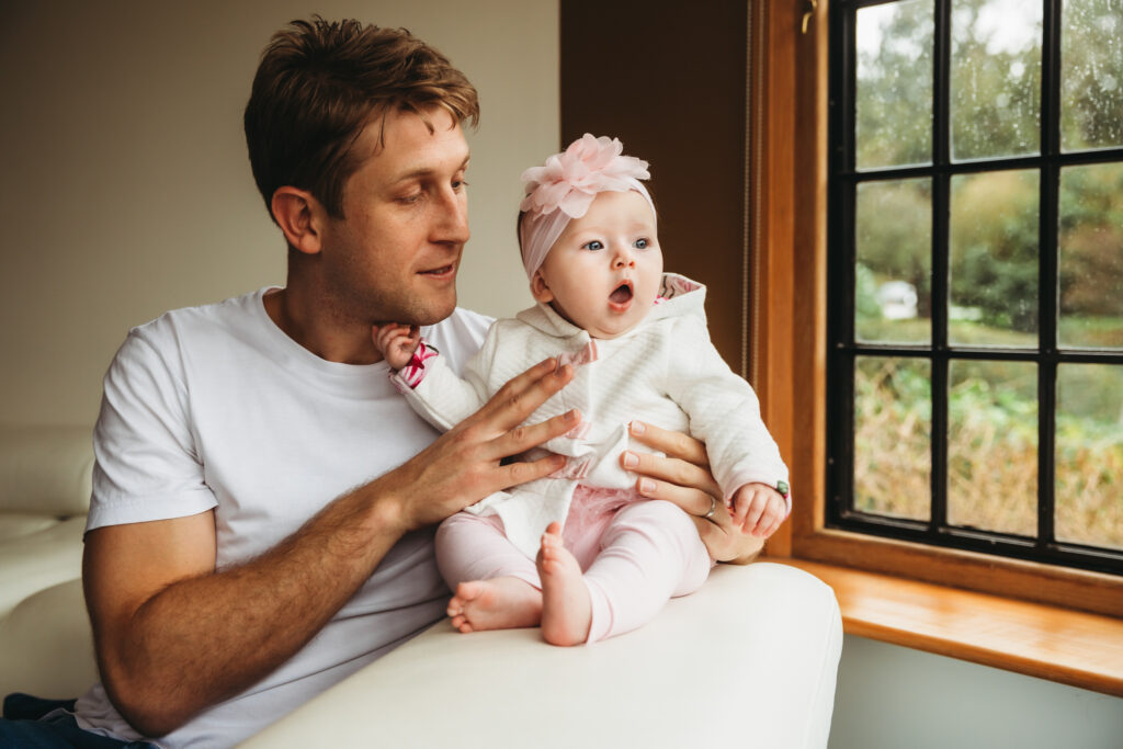 Dad holding his baby daughter on the back of a sofa while she's looking out the window.