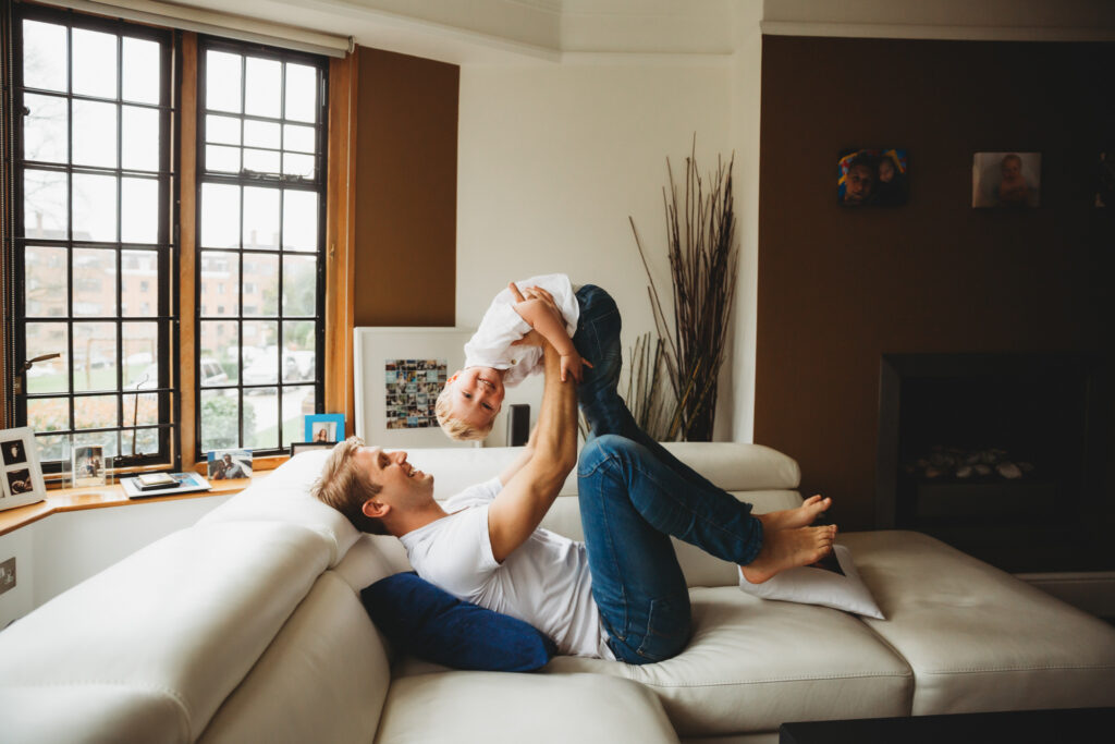 Dad playing with his boy in the lounge lifting him up while boy is looking at the camera.