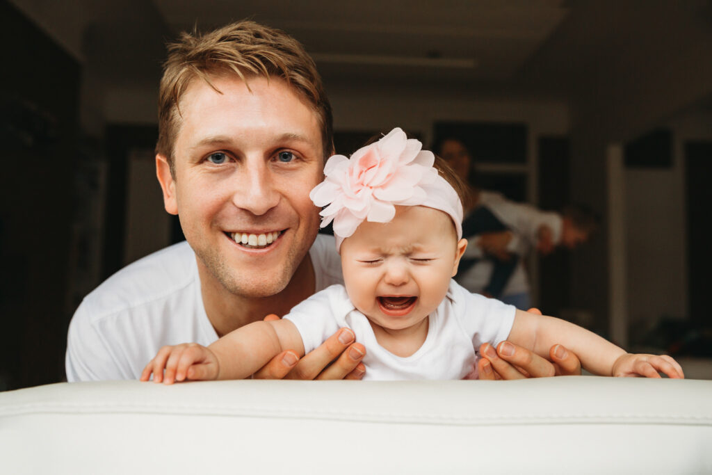 Little baby girl crying while dad is smiling looking at the camera.