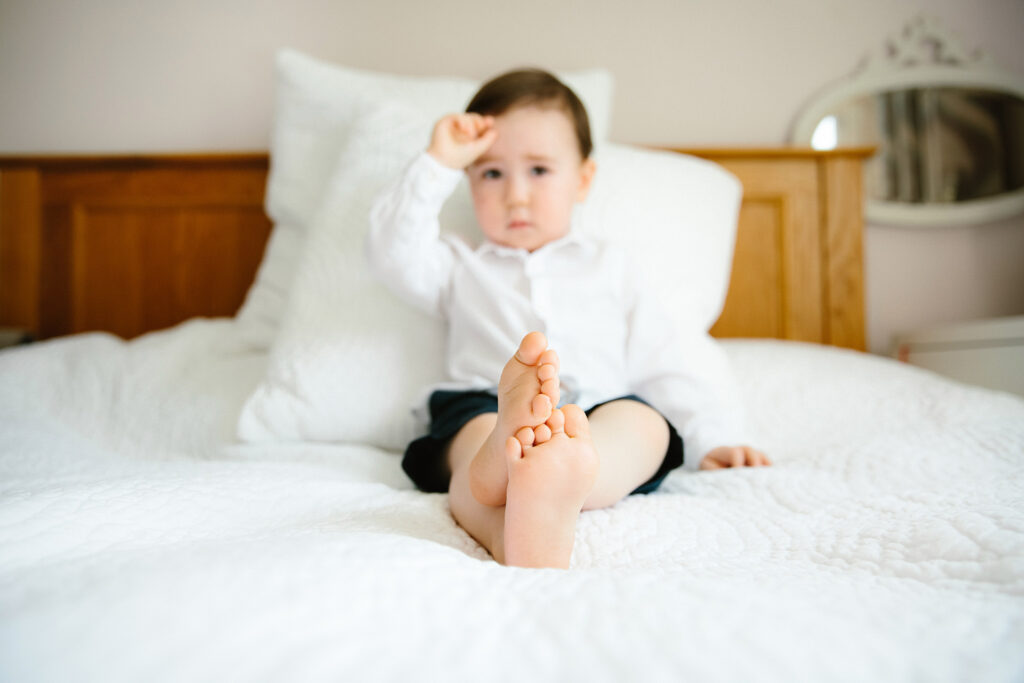 Toddler sitting on bed looking at the camera. His feet are in focus.