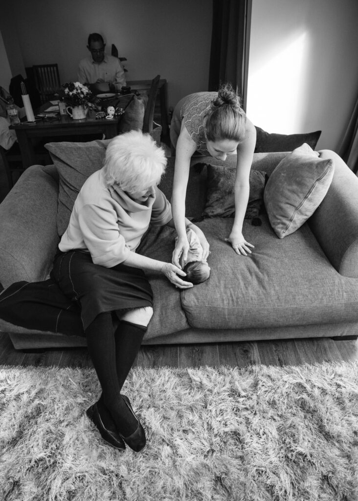 Domumentary style, black and white photo of a baby lying on a sofa whit mum and grandma are looking after him. Dad is working in the background.