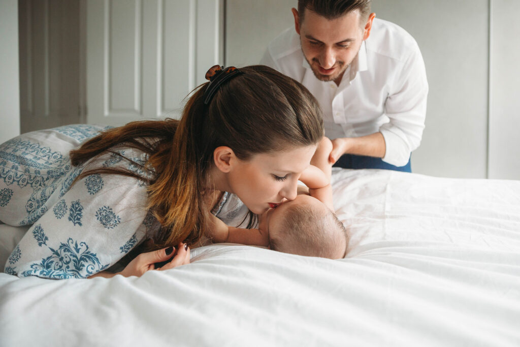 Mum is kissing newborn baby on his forehead on bed by the window.