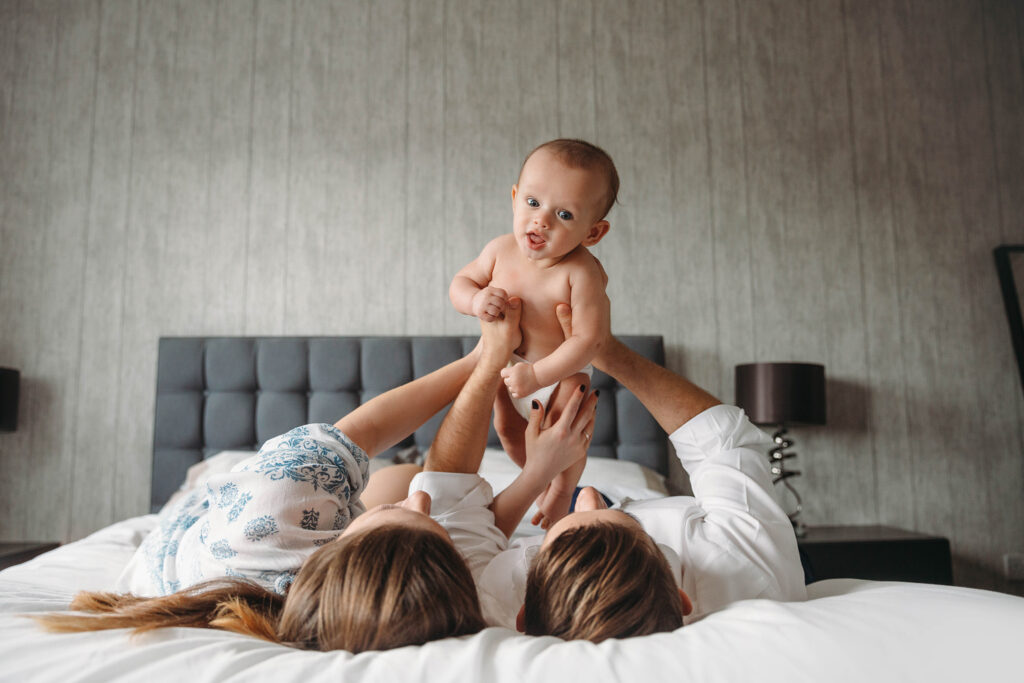 Mum and dad holding their baby up while they are lying down in bed in their home.