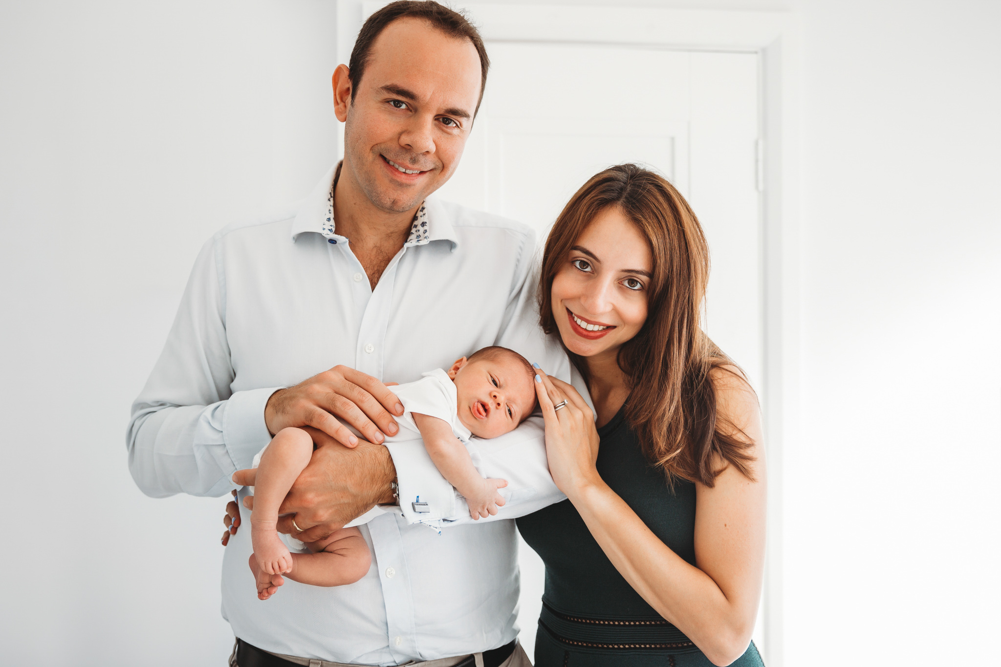 Mum and dad next to each other holding their newborn baby and smiling at the camera.