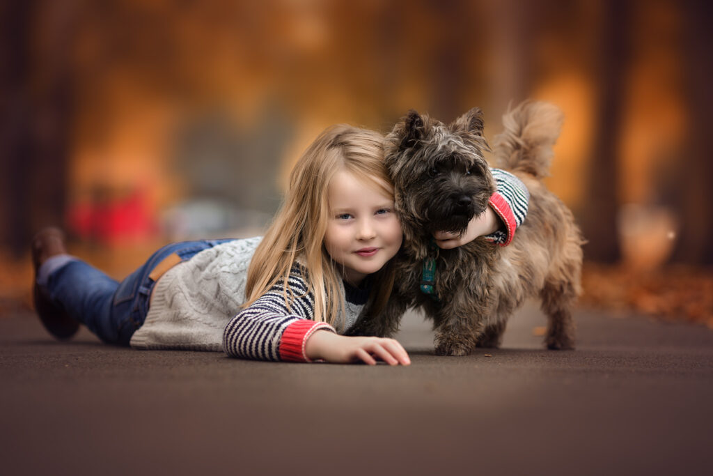 Little girl lying on her tummy on autmn leaves holding her dog and looking at the camera at a park.