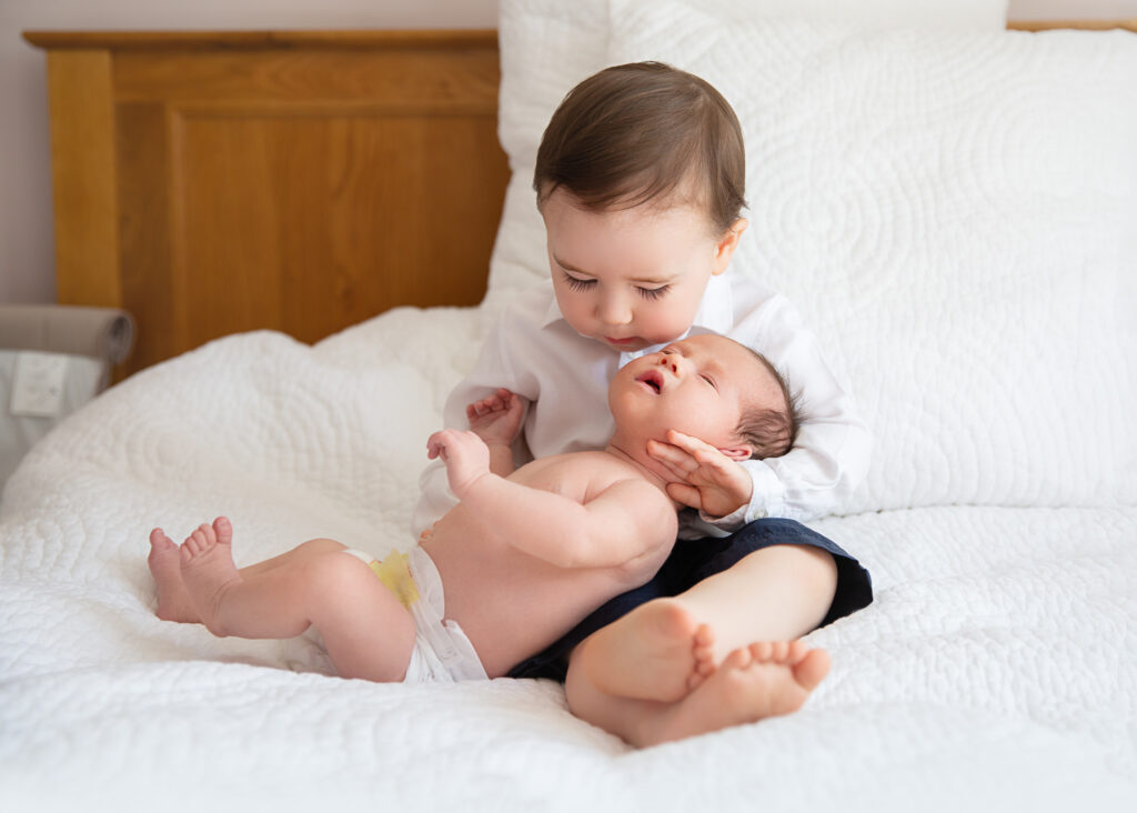 Little boy sitting on bed holding his little baby brother and looking at him with love.