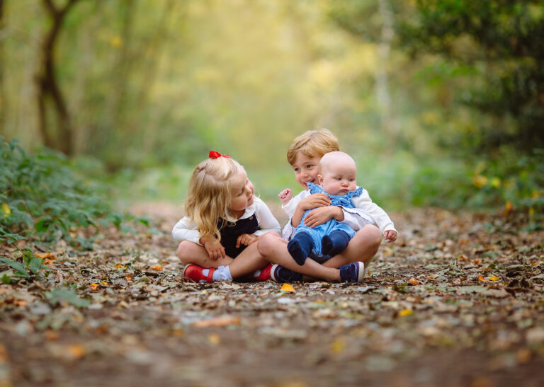 A brother and a sister sitting down at a park holding their baby brother.