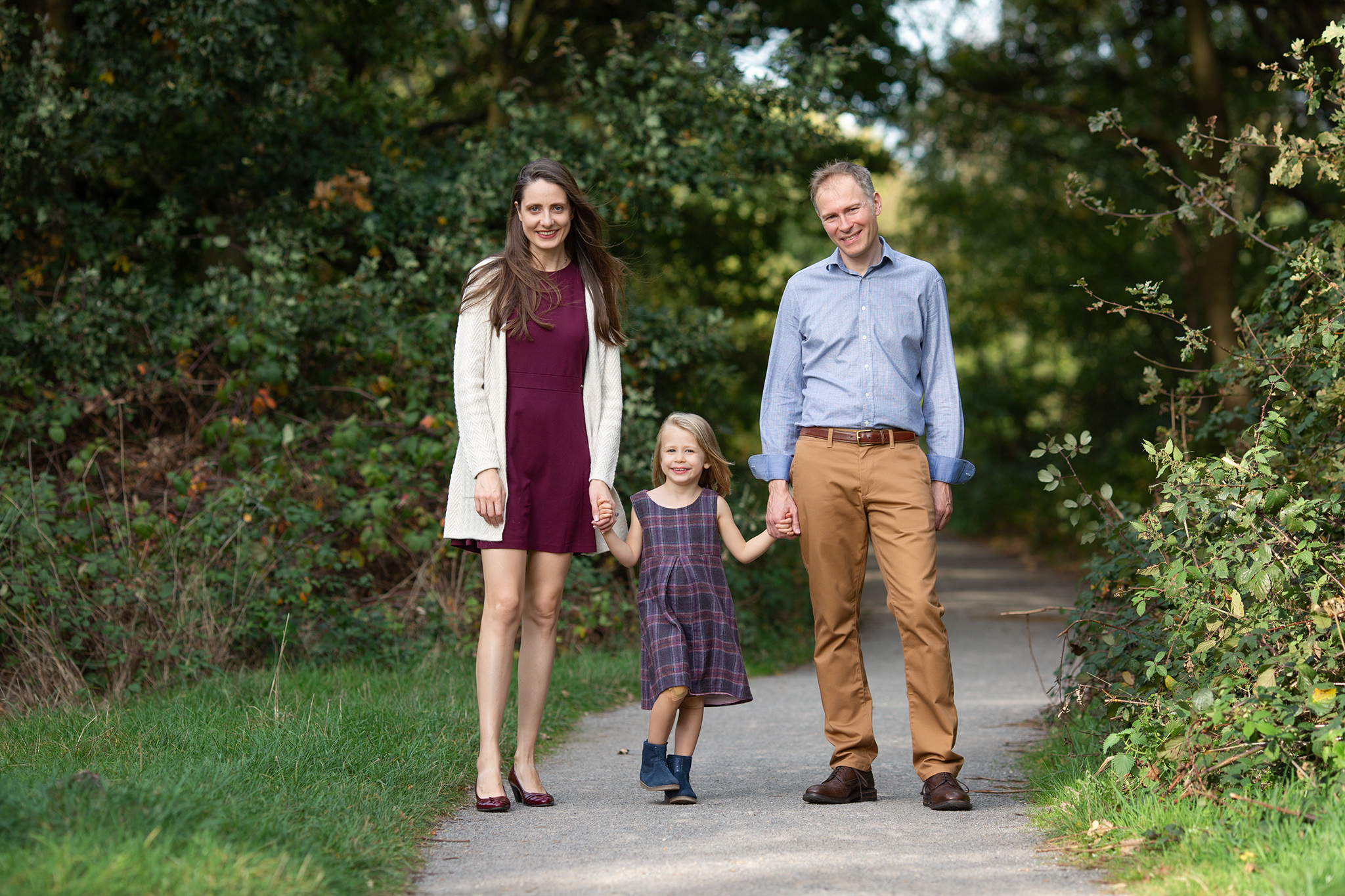 Mum and dad holding their little girl in between them and walking at park smiling at the camera.
