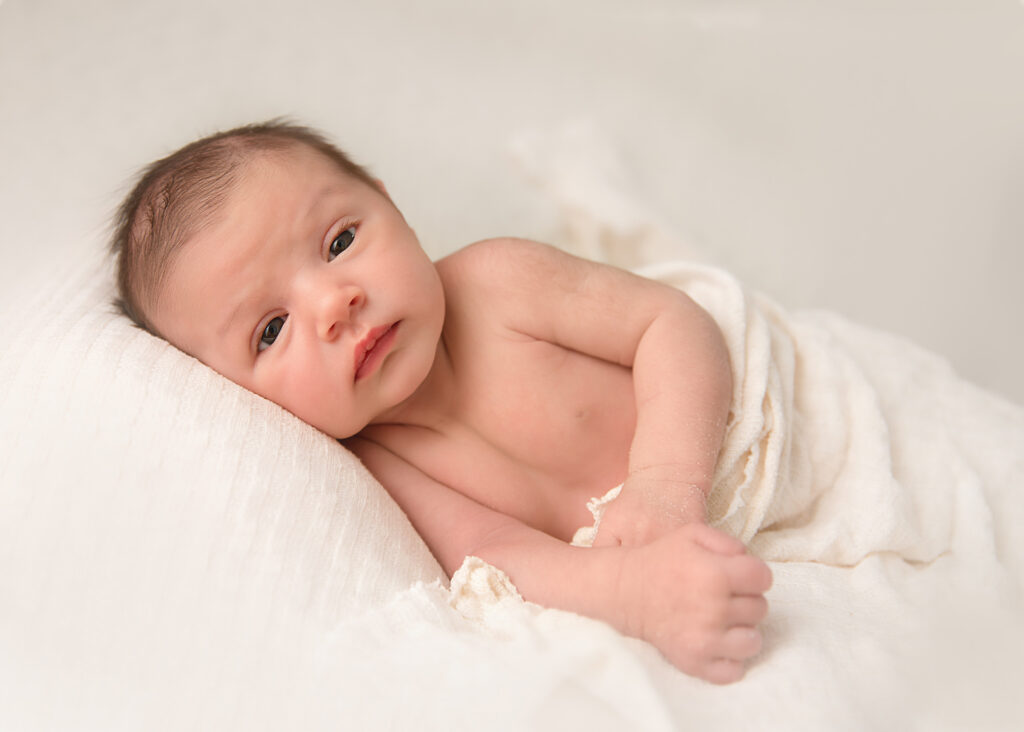Newborn baby lying on her side in cream blanket and sheets looking at the camera.