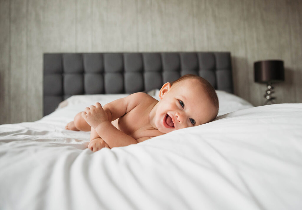 A baby lying in bed in his nappies and smiling.