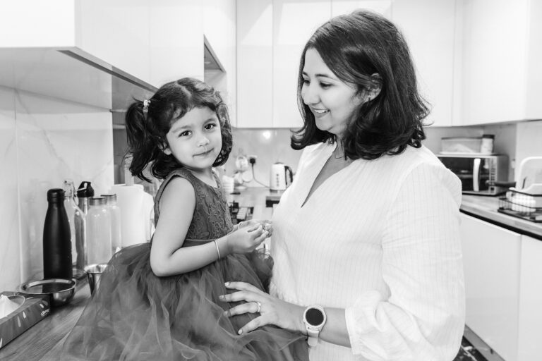 A little girl sitting on the kitchen bench looking and smiling at the camera while her mum is holding her and looking at her.