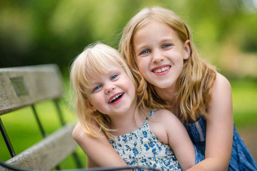 Two sisters sitting on a bench in a park,hugging and smiling at the camera.
