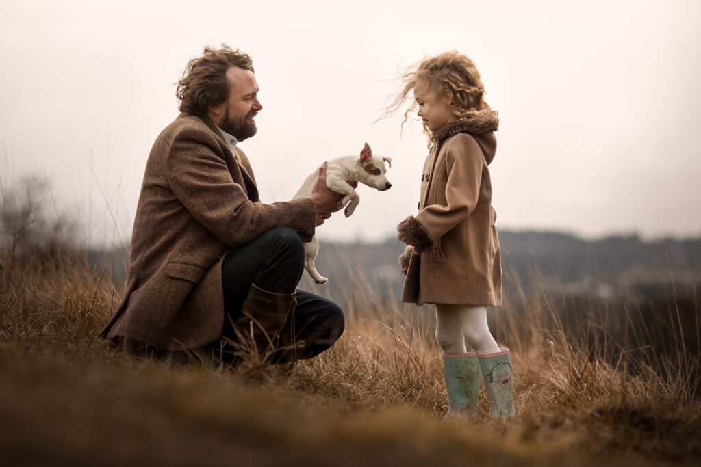 A dad giving his girl a little puppy. They are both on a hill with beautiful views of Surrey.