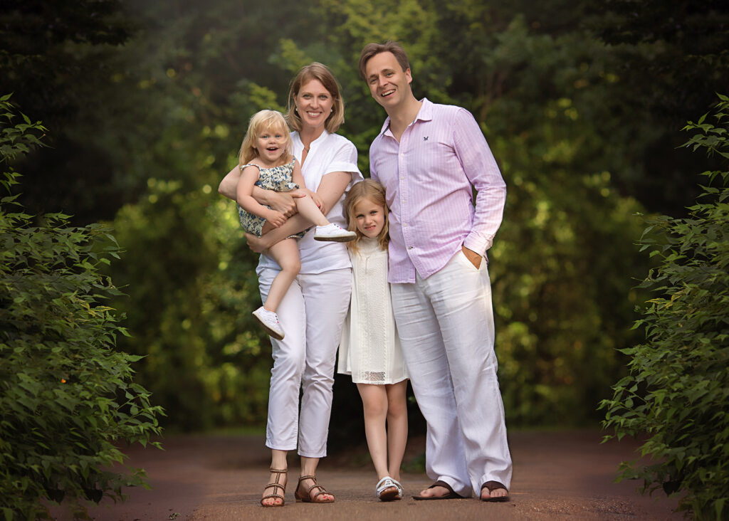Mum, dad and their 2 girls posing for a photo in front of some bushes in a park in London.