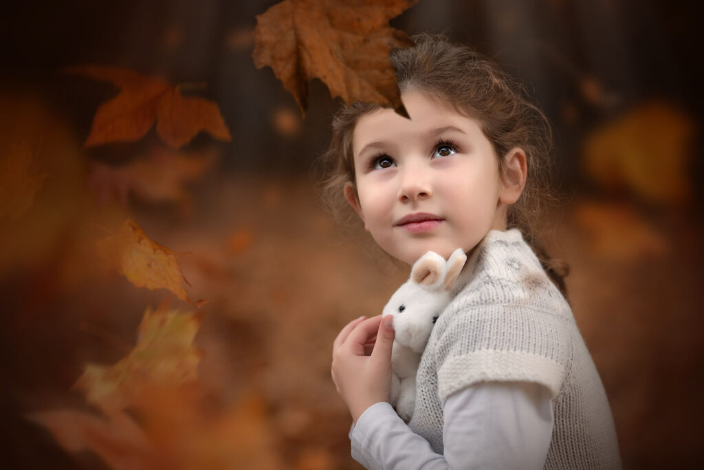 A little girl holding her teddy and looking at falling autumn leaves in Wandsworth Common.