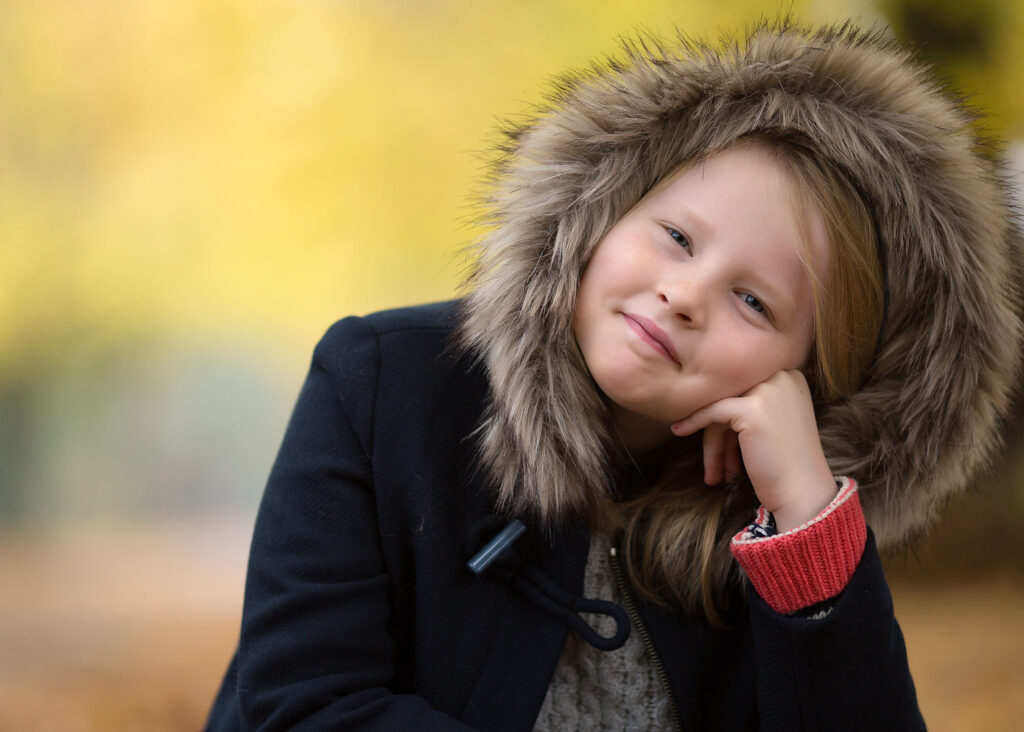 A blond girl wearing a coat and a hoodie sitting down and supporting her head with her hand, posing for a photo at Clapham Common.