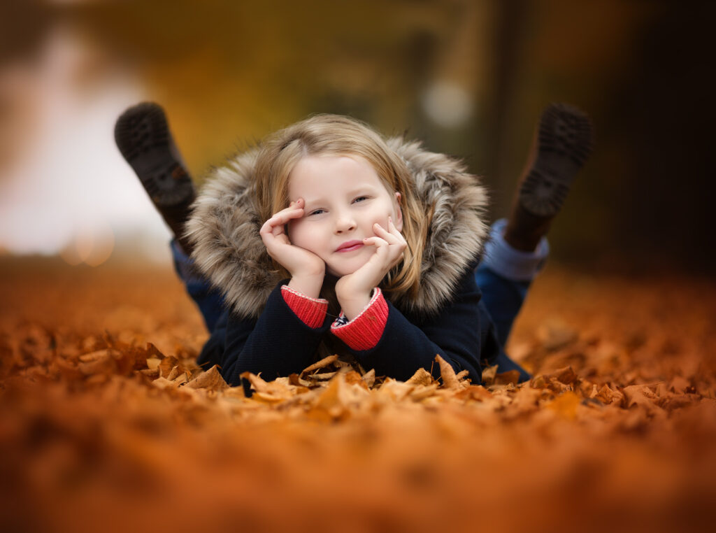 Little girl lying on her tummy on autmn leaves holding her head and looking at the camera at a park.