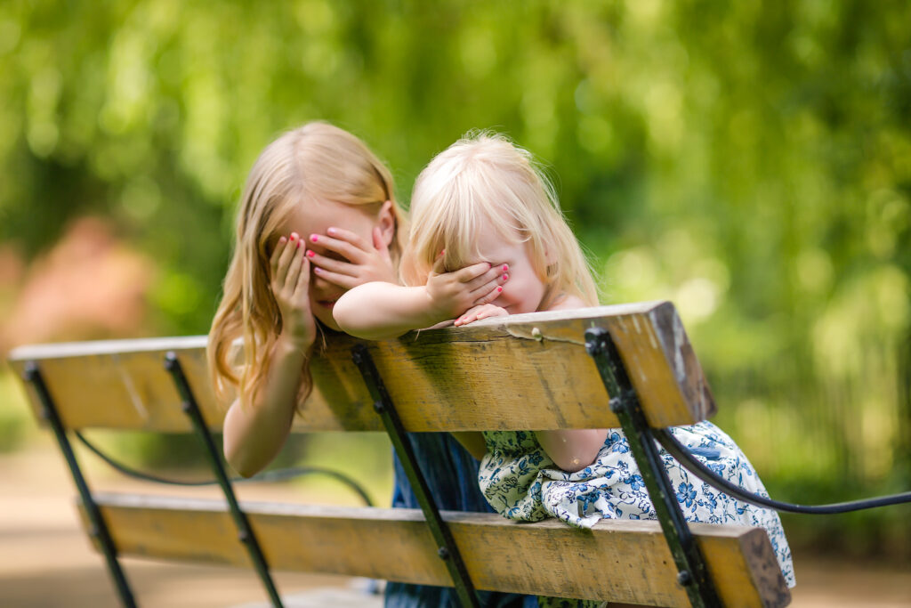 Two little girl sisters sitting on a bench putting their hands in front of of their eyes as if they are playing hide and seek seek in a park in London.