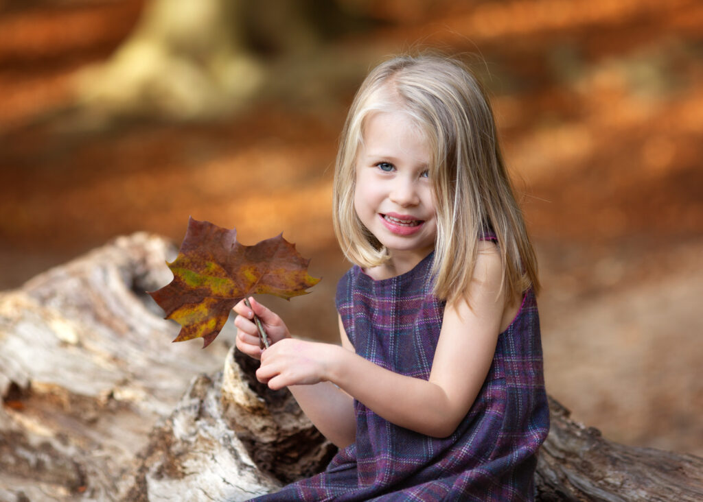 A little girl smiling at the camera holding an autumn leaf for a photo at a park in London.